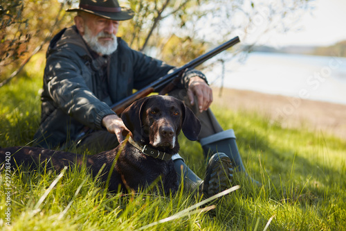 Hunter male with gun sit with faithful dog during hunting. Forest background