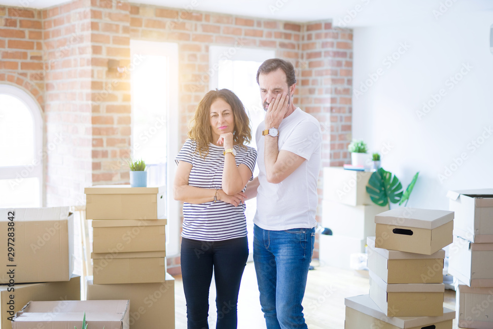 Middle age senior couple moving to a new home with boxes around thinking looking tired and bored with depression problems with crossed arms.