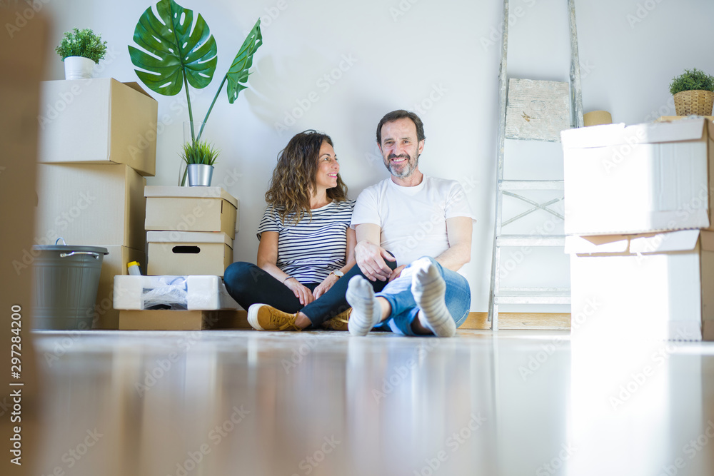 Fototapeta premium Middle age senior romantic couple in love sitting on the apartment floor with cardboard boxes around and smiling happy for moving to a new home
