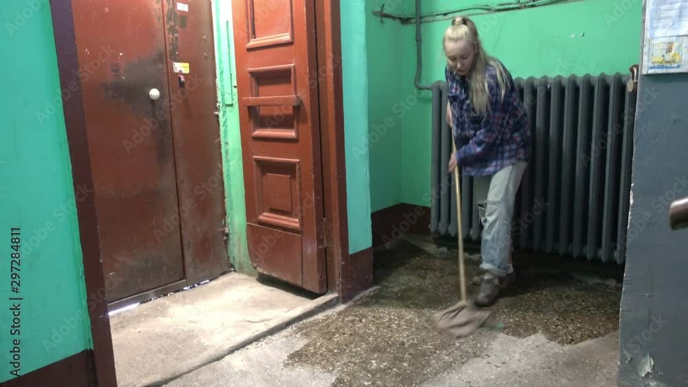 A young woman in blue jeans and a cage shirt swings and washes the floor on a staircase next to an elevator in a Stalinist-built apartment building, Cleaning public spaces by a labor immigrant cleane A young woman in blue jeans and a cage shirt swings and washes the floor on a staircase next to an elevator in a Stalinist-built apartment building, Cleaning public spaces by a labor immigrant cleane