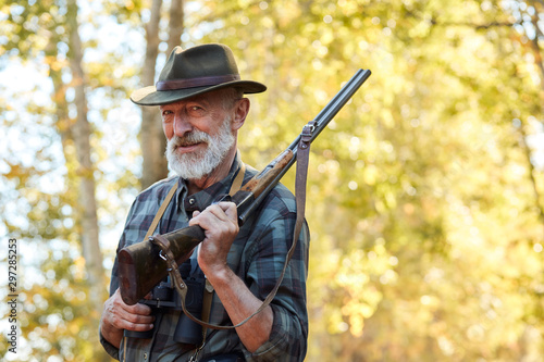 Happy senior hunter smile after getting good, big trophy in forest. Hunting on wild animals, birds. Satisfied hunter with grey beard, holding shotgun on shoulder © alfa27