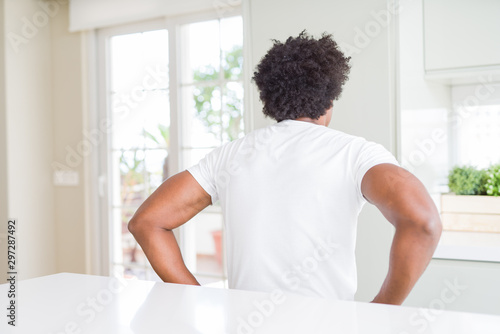 Young african american man wearing casual white t-shirt sitting at home standing backwards looking away with arms on body