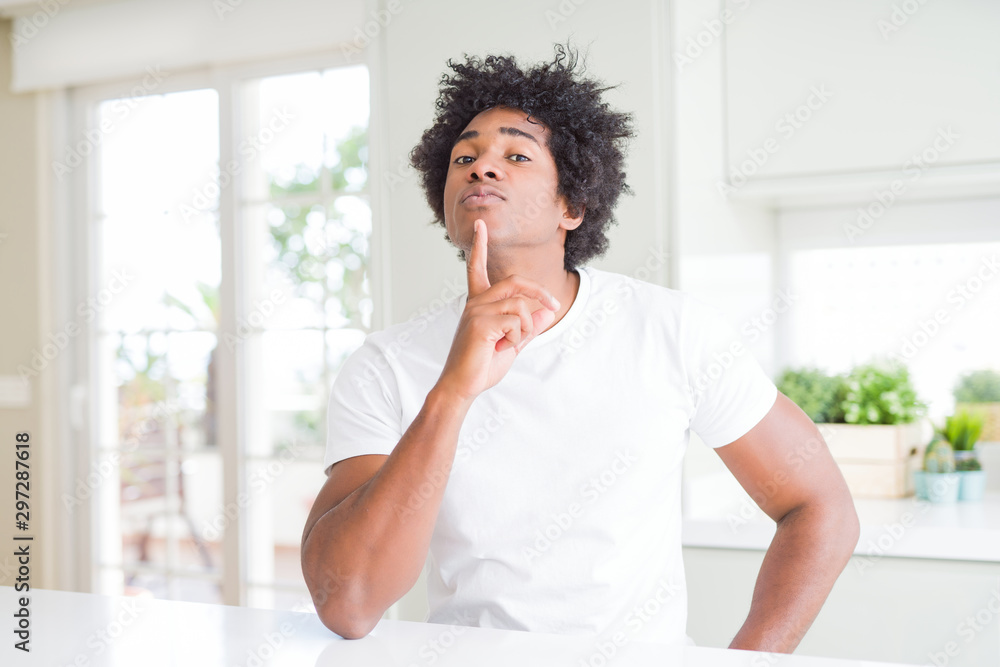 Young african american man wearing casual white t-shirt sitting at home Thinking concentrated about doubt with finger on chin and looking up wondering Young african american man wearing casual white t-shirt sitting at home Thinking concentrated about doubt with finger on chin and looking up wondering