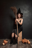 Image of scary witch girl in hat sitting with broom by halloween pumpkins