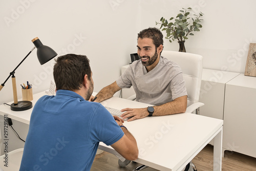 Dental doctor, meeting in his office and sitting at a table, attends with a smile to a client wearing a blue polo shirt in a modern dental clinic.