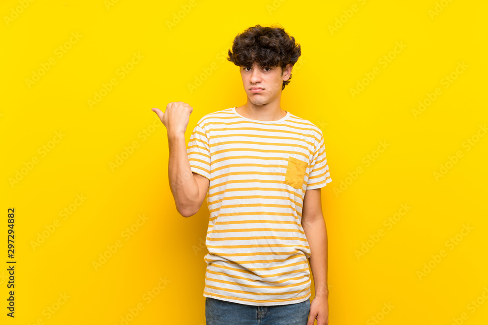Young man over isolated yellow wall unhappy and pointing to the side Young man over isolated yellow wall unhappy and pointing to the side