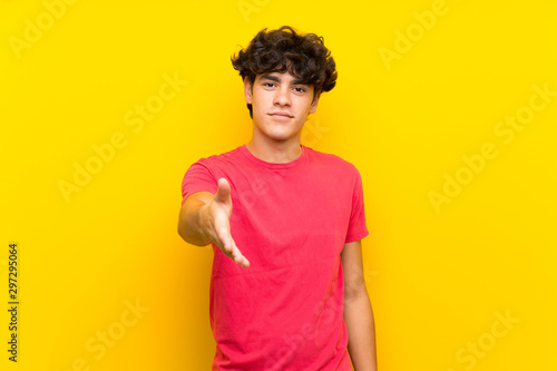 Young man over isolated yellow wall handshaking after good deal