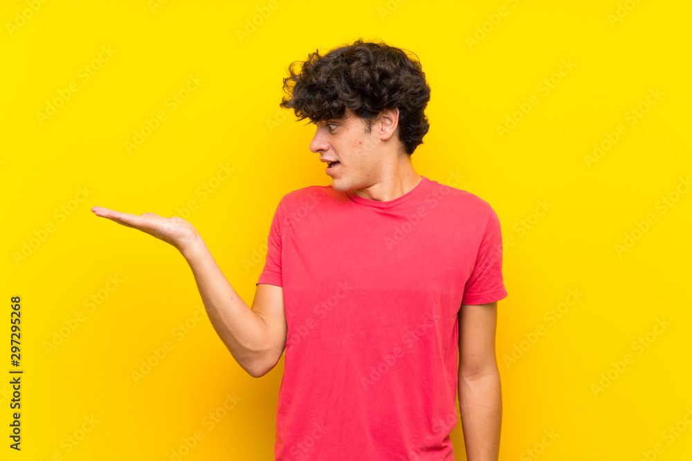 Young man over isolated yellow wall holding copyspace imaginary on the palm Young man over isolated yellow wall holding copyspace imaginary on the palm