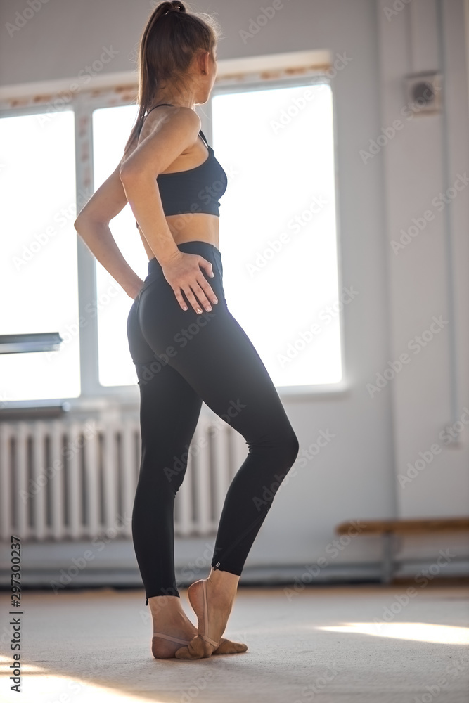Graceful sporty woman in good shape and with long hair, stands with back towards camera, looks dreamy at window, puts hands on hips, stretches fingers on legs, professional sport concept Graceful sporty woman in good shape and with long hair, stands with back towards camera, looks dreamy at window, puts hands on hips, stretches fingers on legs, professional sport concept
