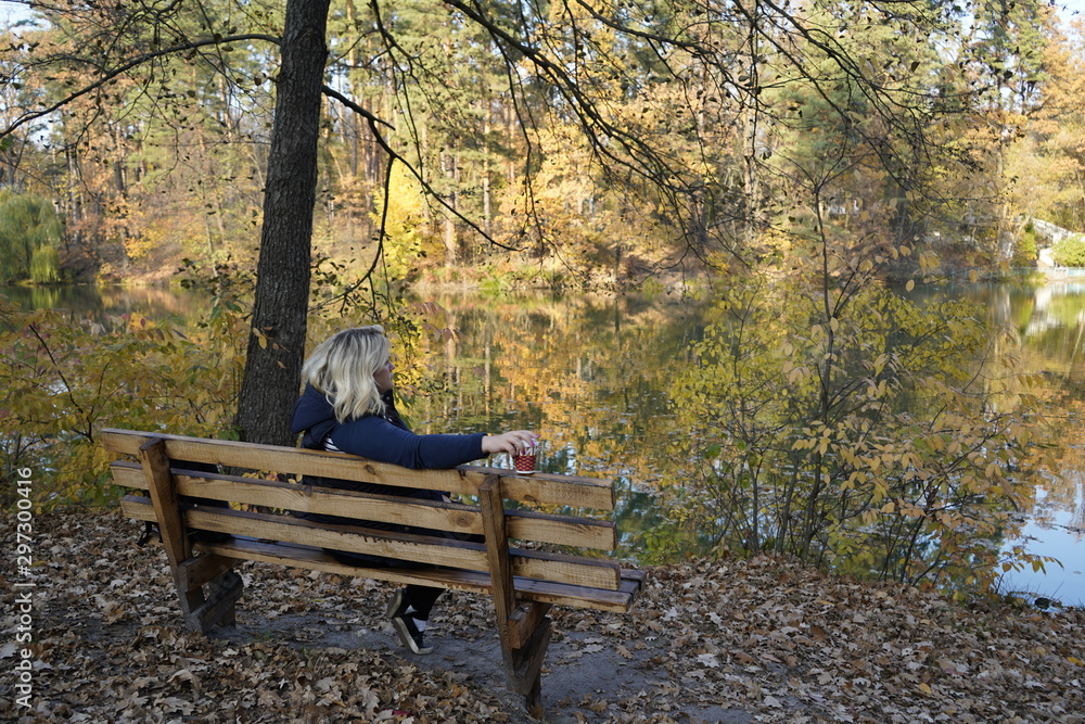 A girl sits on a bench against the background of a lake and autumn forest and drinks coffee in a glass. The girl is resting on a bench in the autumn forest in front of the lake and drinking coffee. A girl sits on a bench against the background of a lake and autumn forest and drinks coffee in a glass. The girl is resting on a bench in the autumn forest in front of the lake and drinking coffee.