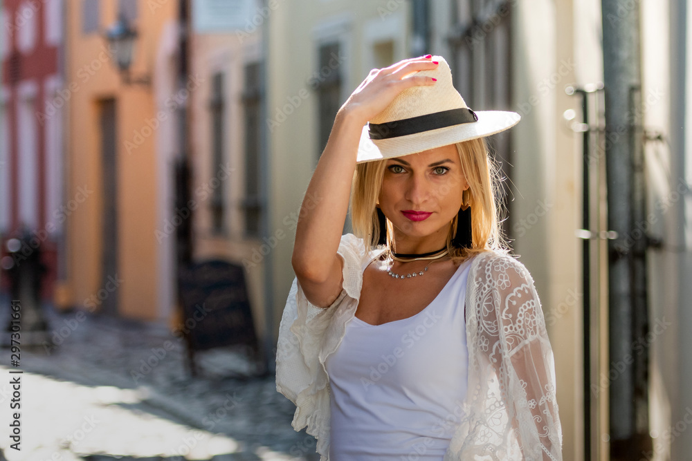 Portrait of beautiful blond woman with sun hat dressed in  light clothes. Trendy girl posing in the street background - image