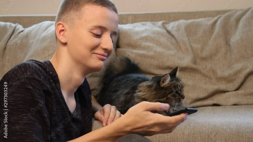 Young teenager boy smiling while watching video on his cell phone sitting near his pet, cat looking at smartphone Young teenager boy smiling while watching video on his cell phone sitting near his pet, cat looking at smartphone