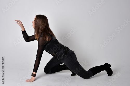 full length portrait of a pretty brunette woman wearing black leather fantasy costume. Sitting down with  a studio background.