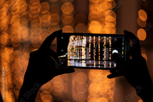 Woman Clicking Photos With Her Smartphone. Raised hand taking wonderful blurred bright yellow orange bokeh background images of festive season Diwali, Christmas, Happy New Year with space for text.