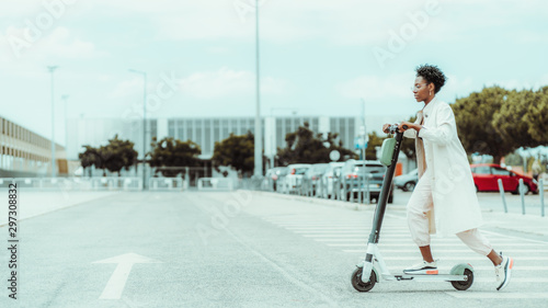 Young fancy African woman in spectacles and a white cloak is riding an e-scooter with green battery above on the asphalt road with a painted arrow as a road marking, sunny day, shallow depth of field © skyNext