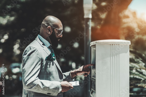 A mature bald black man entrepreneur with a beard and in spectacles is using a parking pay station outdoors; an African businessman is putting his car number into the parking payment terminal