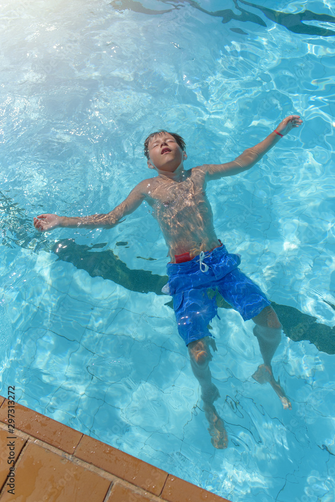 Boy relaxing at hotel swimming pool in summer. He is laying on his back with open hand and legs and closed eyes.