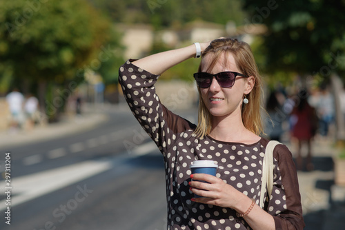 Portrait of a young blondie woman walking in European city and drinking coffee from paper mug, she is smiling to the camera and enjoying her coffee break. © Artem