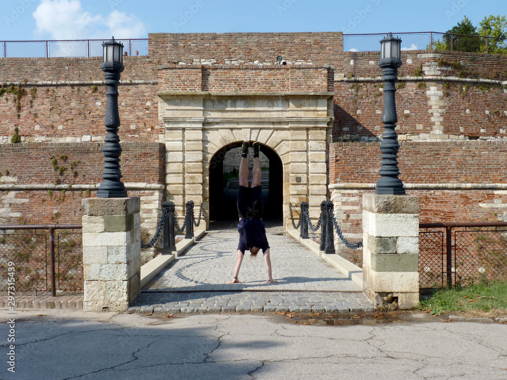 Boy trying to handstand on an old stone bridge between two street lamps, brick fortress in the background
