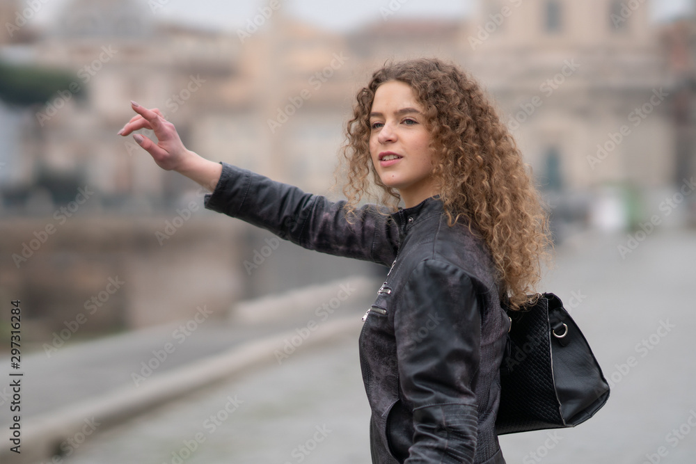 Attractive woman with curly hair and black leather jacket searching for a taxi in Rome, Italy