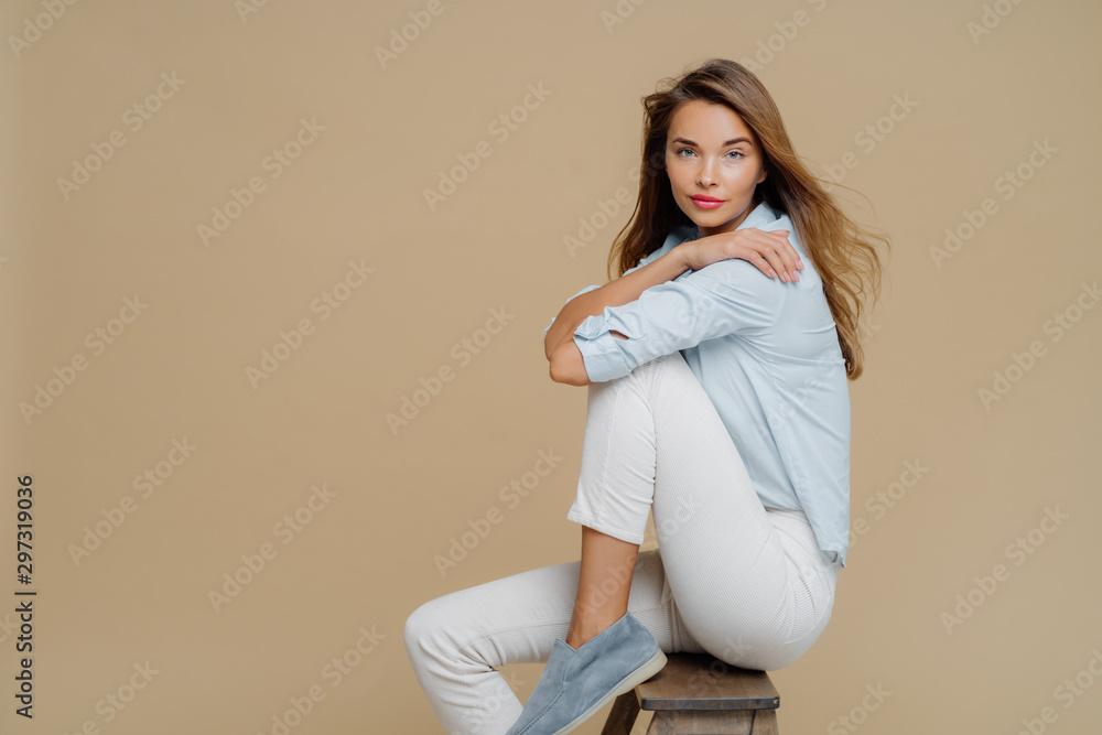 Studio shot of restful beautiful Caucasian woman sits on chair, wears shirt, white trousers and shoes, keeps hands crossed over body, has confident look at camera, isolated over brown background. Studio shot of restful beautiful Caucasian woman sits on chair, wears shirt, white trousers and shoes, keeps hands crossed over body, has confident look at camera, isolated over brown background.