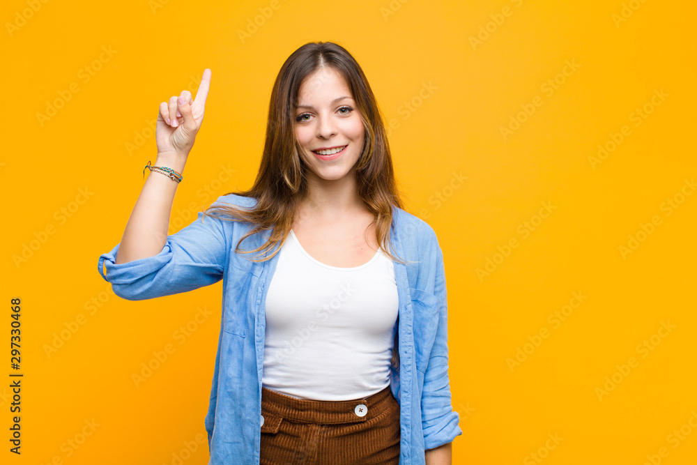 young pretty woman smiling cheerfully and happily, pointing upwards with one hand to copy space against orange wall - obrazy, fototapety, plakaty 