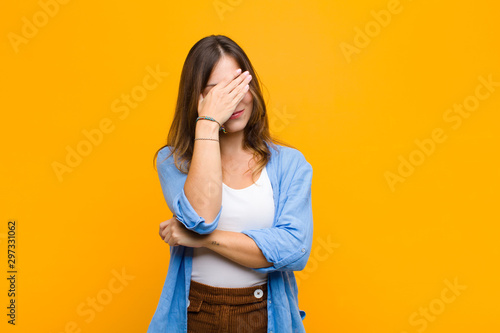 young pretty woman looking stressed, ashamed or upset, with a headache, covering face with hand against orange wall