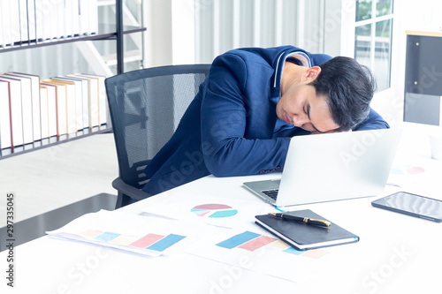 A young businessman sitting in a modern office. He has a feel sleepy because  hard work so tired weary fatigued and exhausted. On his table have a computer laptop tablet pen paper graph. photo