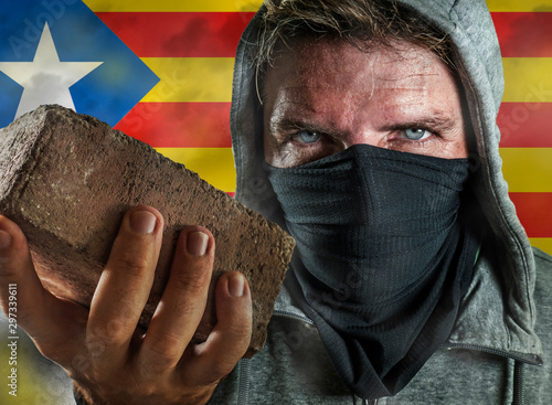 young Catalan man as ultra and radical separatist holding brick protesting in face mask holding brick threatening at nationalistic riot isolated on Catalonia independentist flag