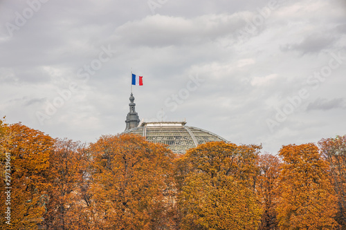 French flag waving on the roof of the Grand Palais in Paris on a fall day surrounded by a giant statue and by colored trees with autumn leaves