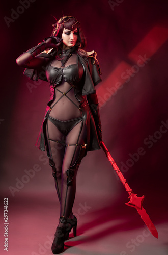 Beautiful leggy busty cosplay girl wearing an erotic leather costume posing with a fake spear on a red background in theatrical smoke.