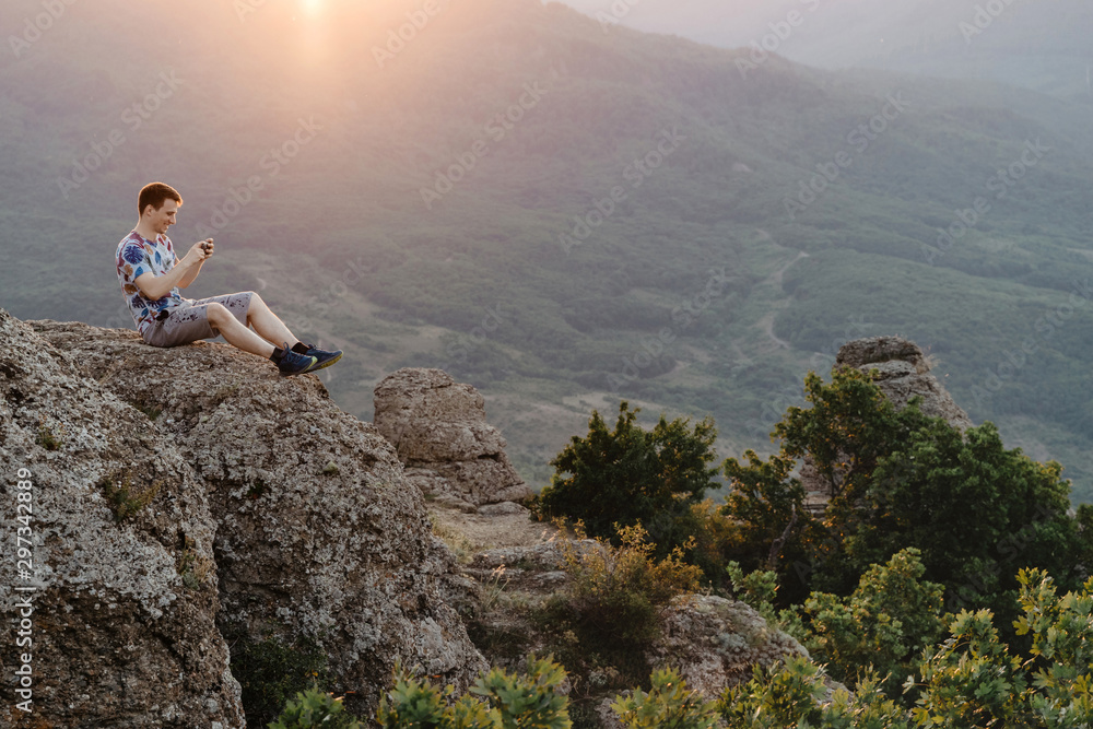 Young man sits on a cliff and using a smartphone in mountains during sunset.