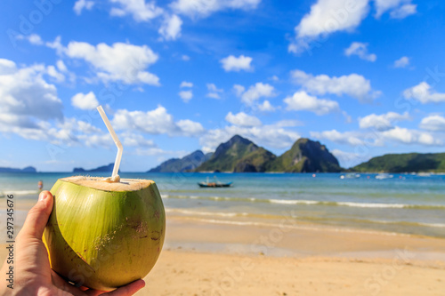 Hand holding a coconut coco loco coctail with straw, tropical islands, beach, sea and blue sky in the background, El Nido, Palawan, Phillipines