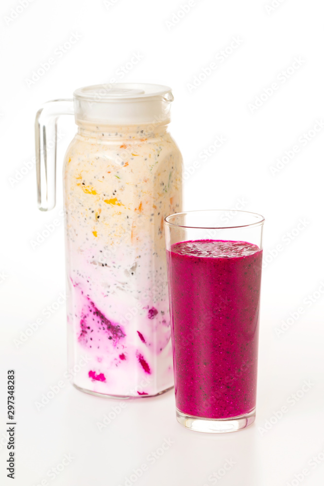 Jug of sweet colorful layered yougurt with fruits and berries and a glass of dragon fruit juice at white background. Concept of detox and vegan healthy lifestyle. Jug of sweet colorful layered yougurt with fruits and berries and a glass of dragon fruit juice at white background. Concept of detox and vegan healthy lifestyle.