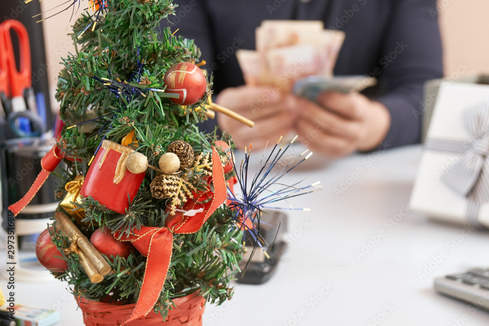 Decotated small Christmas tree standing on the table on the foreground and a manager getting a bonus money on workplace in Christmas or New Year eve on the background