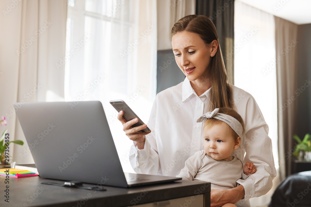 Woman holding smartphone in hand. Little girl sitting on mother's knees and watching cartoons online at laptop. Female sending messages and commenting in social networks, looking at screen of mobile Woman holding smartphone in hand. Little girl sitting on mother's knees and watching cartoons online at laptop. Female sending messages and commenting in social networks, looking at screen of mobile
