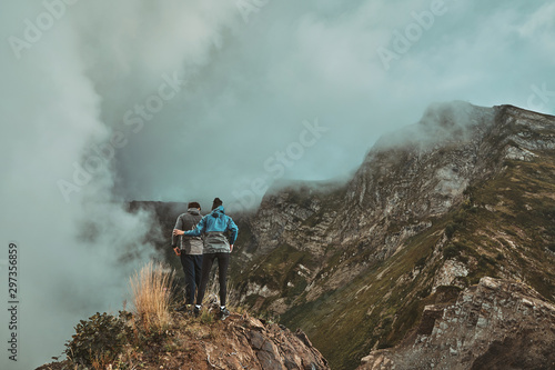 a young couple stands on a cliff in the mountains and looks down against the background of a mountain top with clouds