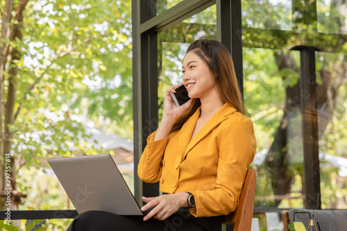 Business Asian young woman working with laptop smile and talking with customer in smartphone to get idea and requirement for success job outside office in coffee shop,Small Business Startup Concept