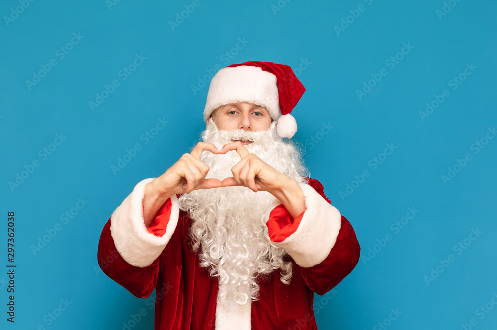 Handsome young Santa Claus shows heart gesture with his hands and looks into the camera with a positive face isolated on a blue background. Santa loves you. Christmas and New Year. Handsome young Santa Claus shows heart gesture with his hands and looks into the camera with a positive face isolated on a blue background. Santa loves you. Christmas and New Year.