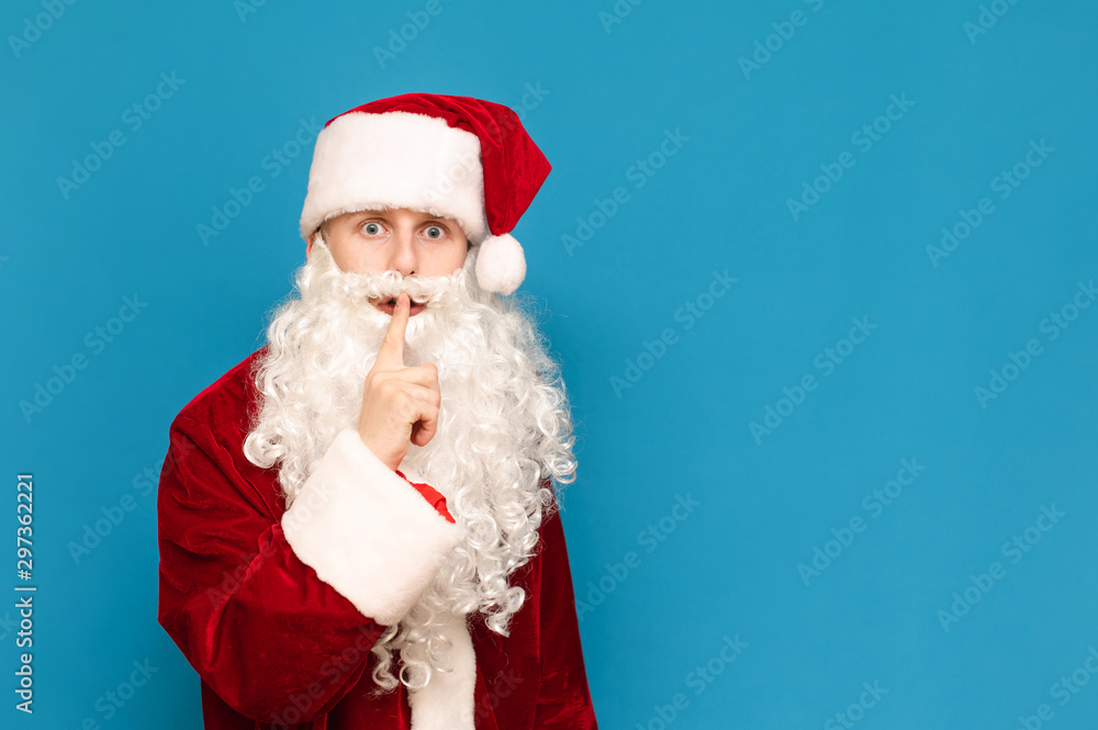 Scared boy in santa costume looks into camera with shocked face and shows gesture of silence with hand on blue background. Young Santa asks for silence, isolated. Tshshsh. Copy space Scared boy in santa costume looks into camera with shocked face and shows gesture of silence with hand on blue background. Young Santa asks for silence, isolated. Tshshsh. Copy space