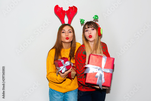 Portrait of two cheery girls dressed in sweaters and christmas hats hugging and looking at camera isolated over white background. Brightfull expressions of happy emotions of two amazing girls friends photo