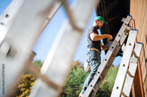 Fototapete A side view of mature man outdoors in summer, painting wooden house