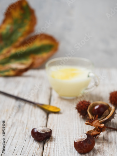 Chestnuts on a wooden surface. In the background is a glass cup with golden milk and a leaf of chestnut. Close-up.