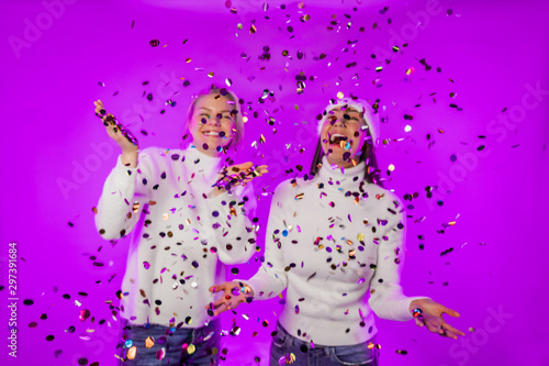  Girls laugh happily. Metallic shiny confetti falls. Christmas mood. Girls in white fluffy sweaters on a bright purple isolated neon background.