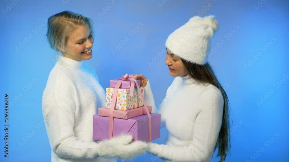 A girl gives a gift to her friend. Girls rejoice and smile at each other. Girls in white fluffy sweaters on a bright blue isolated neon background. Christmas mood. Neon party light. A girl gives a gift to her friend. Girls rejoice and smile at each other. Girls in white fluffy sweaters on a bright blue isolated neon background. Christmas mood. Neon party light.