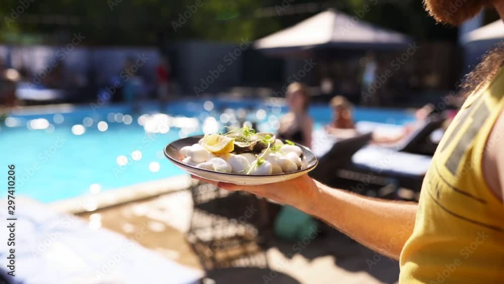 Waiter carries oysters and scallops on tray, with slices of lemon with a swimming pool on background at luxury hotel or beach club. Man serving food in slow motion at resort. Seafood on a plate. Waiter carries oysters and scallops on tray, with slices of lemon with a swimming pool on background at luxury hotel or beach club. Man serving food in slow motion at resort. Seafood on a plate.