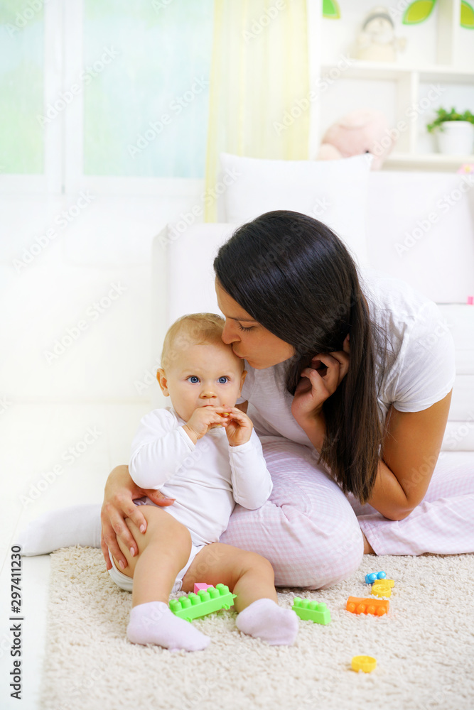 Young mom kissing her baby, sitting on the floor, playing with baby toys Young mom kissing her baby, sitting on the floor, playing with baby toys