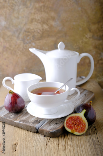 A white cup and a white teapot with tea, a jar of honey and fresh ripe figs on the cutting board on the table.