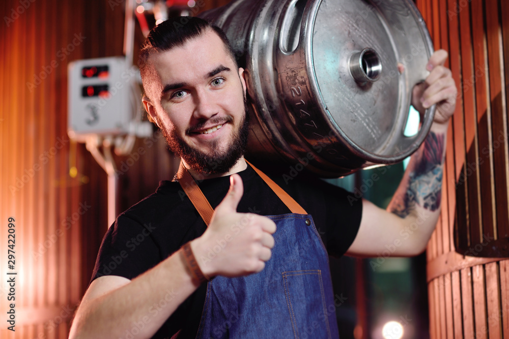 Fototapeta premium a young handsome male brewer holds an iron barrel with beer on the background of the brewery and beer tanks