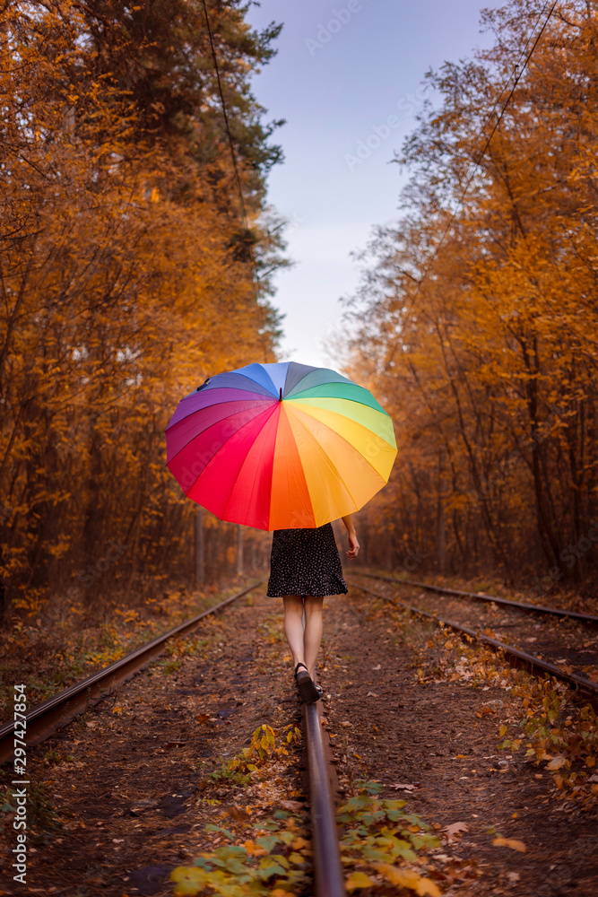 A young girl with a multi-colored umbrella is walking along the tram. Walk through the autumn forest.